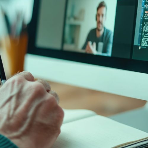 Virtual Learning and Coding Session at Home Workspace. Online Body Doubling. A person takes notes while engaged in a virtual coding class An instructor is visible on the computer screen creating a focused and productive atmosphere in this home office setup